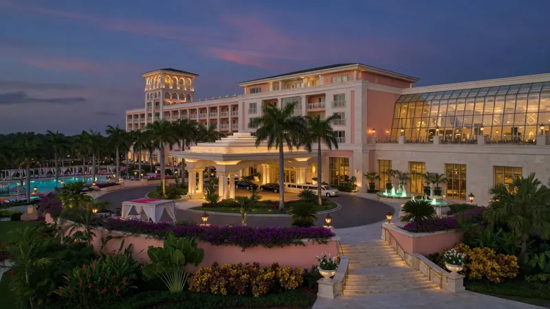 Twilight view of a luxurious pink and white resort hotel with a grand entrance, fountain, palm trees, and illuminated pool area.